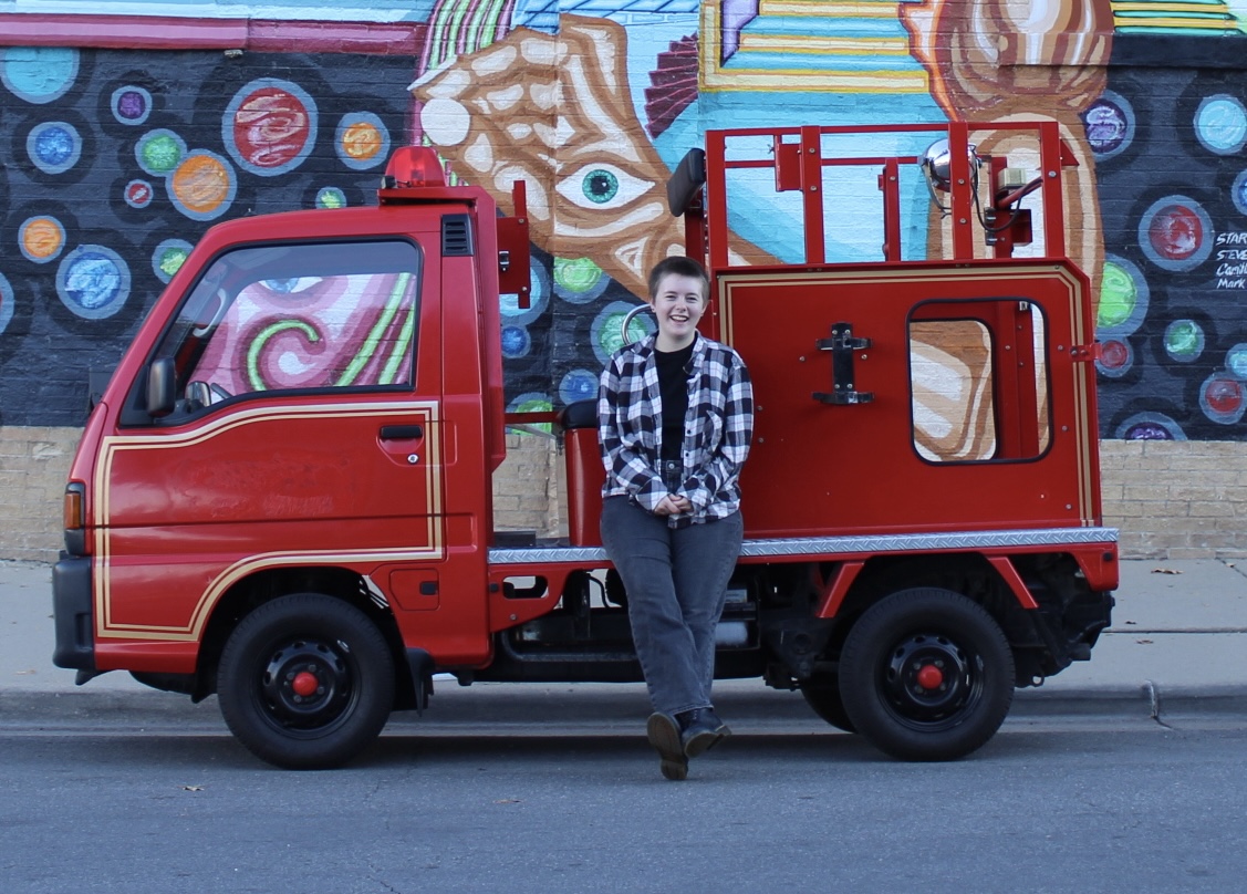 Jordan stands confidently in front of a miniature red fire truck, wearing a plaid shirt and jeans. Behind them, a vibrant street mural features circular patterns and abstract designs.